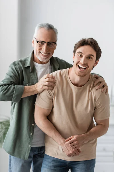 Sonriente hombre en gafas abrazando hombros de hijo excitado mientras mira a la cámara - foto de stock