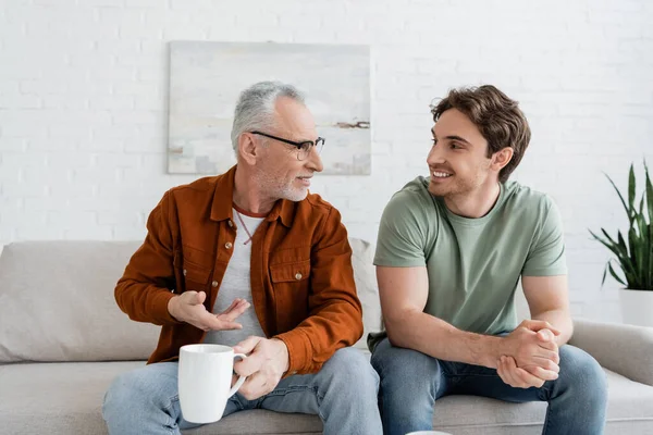 Senior man with tea cup gesturing and talking to young cheerful son on couch at home — Stock Photo