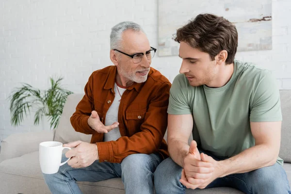 Grey haired man holding tea cup and talking to young son on sofa in living room — Stock Photo