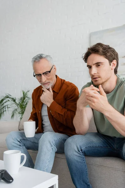 Thoughtful mature man with tea cup touching beard while sitting near serious son — Stock Photo
