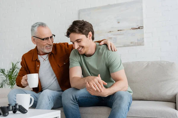 KYIV, UKRAINE - MAY 11, 2022: smiling man with tea cup hugging shoulders of smiling son while talking to him at home — Stock Photo