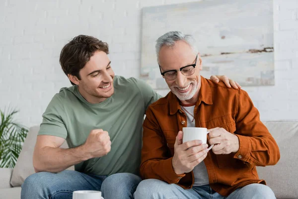 Excited man showing win gesture while talking to smiling dad holding tea cup on couch at home — Stock Photo