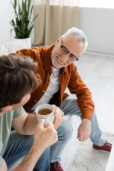 High angle view of mature man in eyeglasses smiling and talking to son with tea cup on blurred foreground — Stock Photo