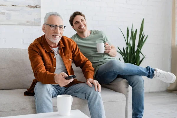 Mature man with remote controller clicking channels while watching tv with cheerful son holding tea cup — Stock Photo