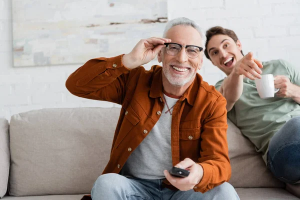 Verschwommener Mann mit Teetasse, der lacht und mit dem Finger zeigt, während er Komödienfilm in der Nähe lächelnden Papas anschaut, der seine Brille justiert — Stockfoto