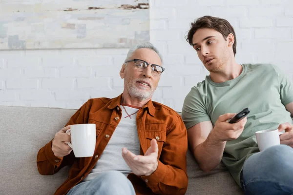 Mature man with tea cup pointing with hand near son clicking tv channels on remote controller — Stock Photo