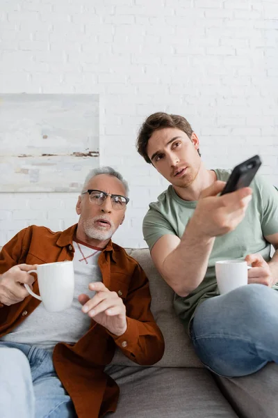 Tensed dad and son with tea cups watching tv on sofa in living room — Stock Photo
