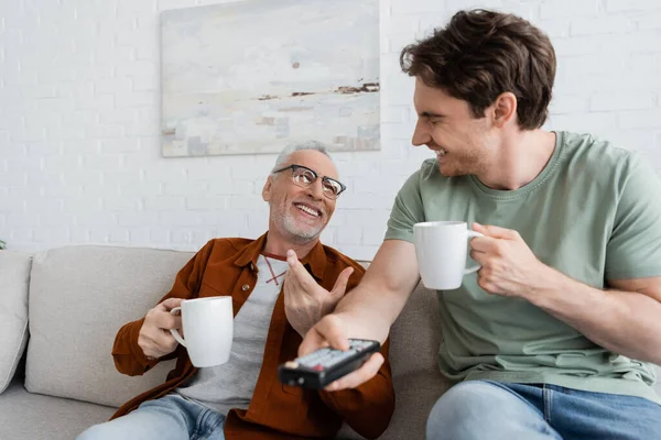 Fröhlicher Mann mit Teetasse im Gespräch mit lachendem Sohn, der Fernsehkanäle auf verschwommenem Vordergrund anklickt — Stockfoto