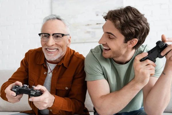 QUIIV, UCRÂNIA - MAIO 11, 2022: homem alegre com joystick olhando para pai sorridente jogando videogame — Fotografia de Stock