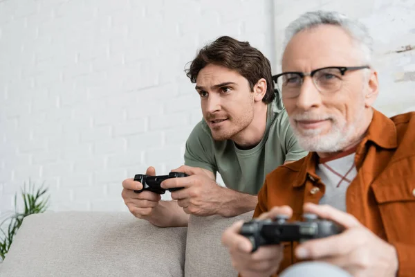 KYIV, UKRAINE - MAY 11, 2022: young and tensed man gaming with mature dad smiling on blurred foreground — Stock Photo