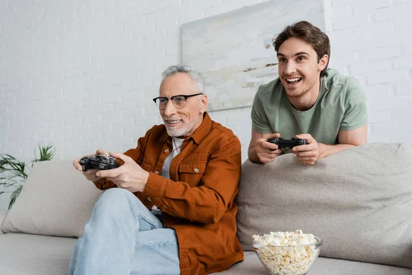 KYIV, UCRANIA - 11 de mayo de 2022: un hombre canoso sonriendo mientras juega un videojuego con su hijo emocionado en el sofá de su casa — Stock Photo