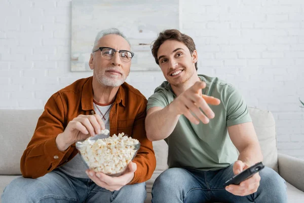 Uomo sorridente che punta con il dito mentre guarda la tv con il padre che tiene in mano ciotola di popcorn — Foto stock