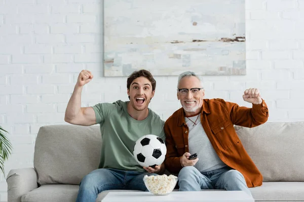 Jeune homme excité avec papa mature montrant geste de victoire près de ballon de football et pop-corn tout en regardant le championnat à la télévision — Photo de stock