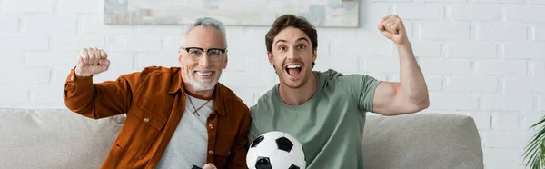 Cheerful father and son with soccer ball showing win gesture while watching football match at home, banner — Stock Photo