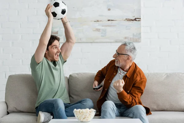 Homem animado segurando bola de futebol em mãos levantadas perto pai sênior mostrando gesto de vitória perto tigela de pipocas — Fotografia de Stock