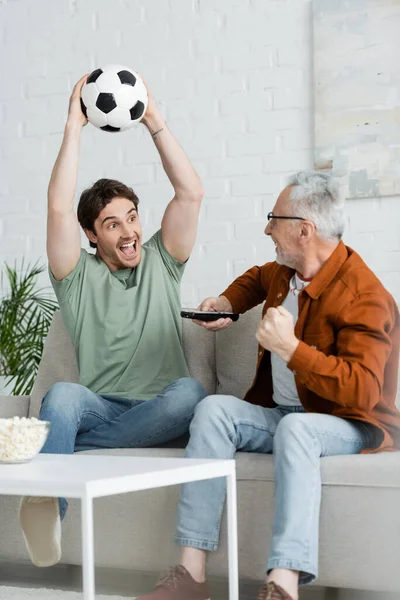 Homem alegre gritando e segurando bola de futebol em mãos levantadas perto de pai maduro mostrando gesto de vitória — Fotografia de Stock