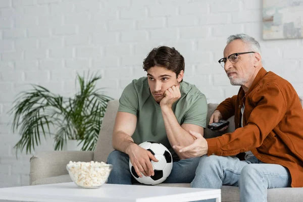 Chateado homem com bola de futebol assistindo campeonato na tv perto pai descontente apontando com a mão — Fotografia de Stock