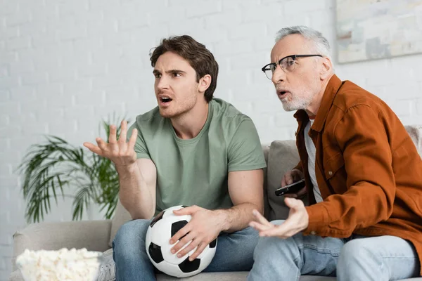 Pai e filho descontentes apontando com as mãos enquanto assiste campeonato de futebol na sala de estar — Fotografia de Stock