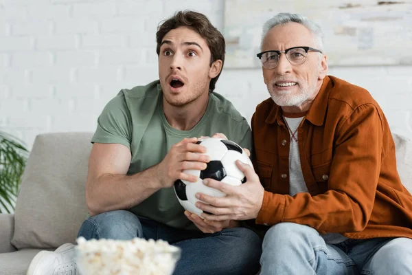 Cara emocionado com pai tenso segurando bola de futebol enquanto assiste campeonato de futebol na tv — Fotografia de Stock