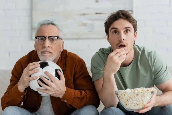 Tenso maduro homem segurando bola de futebol perto filho comer pipocas enquanto assiste campeonato na tv — Fotografia de Stock