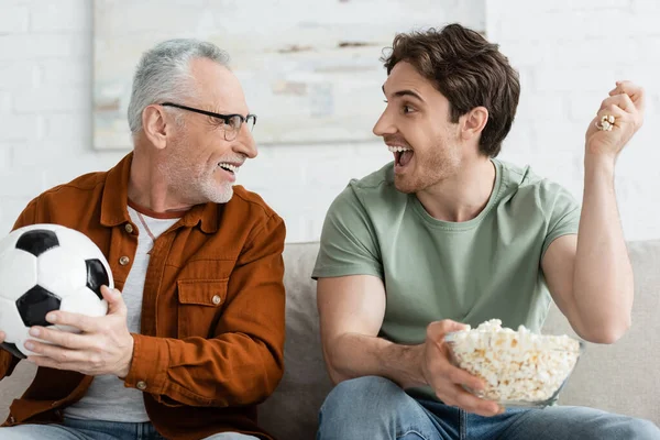 Hombre emocionado con palomitas de maíz y padre feliz con pelota de fútbol mirándose en la sala de estar - foto de stock