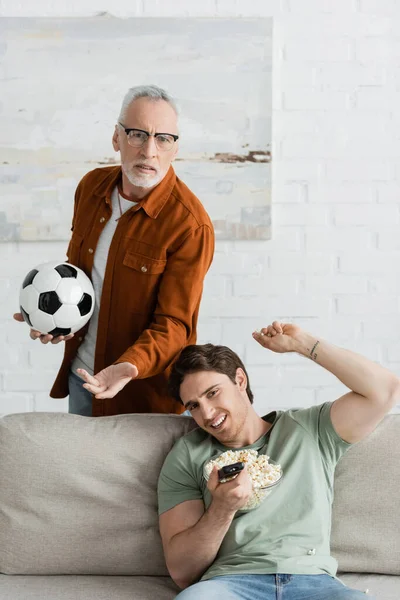 Frustrierte Vater und Sohn mit Fußball und Schale Popcorn gestikulieren, während Fußballmeisterschaft im Fernsehen sehen — Stockfoto