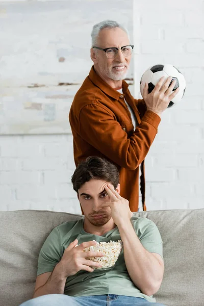 Joven disgustado hombre con palomitas de maíz tocando la frente mientras mira partido cerca de papá maduro con pelota de fútbol - foto de stock