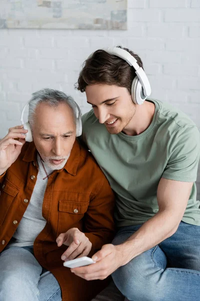 Grey haired man in wireless headphones pointing at smartphone near young and smiling son — Stock Photo