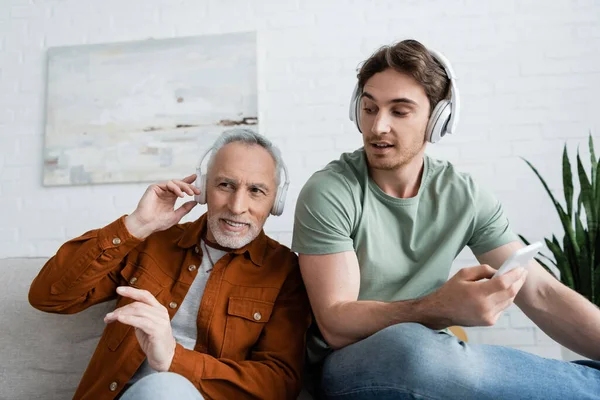 Jeune homme avec téléphone portable regardant papa mature souriant tout en écoutant de la musique dans les écouteurs — Photo de stock