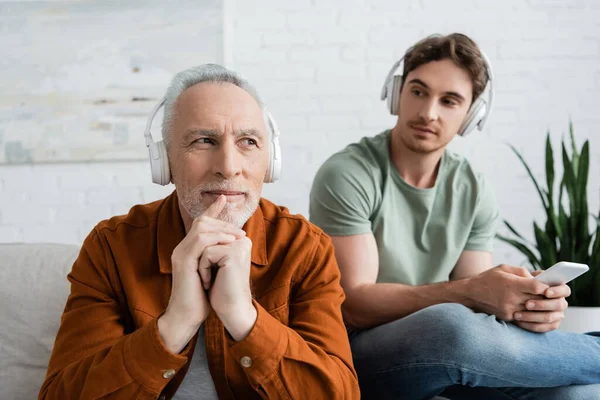 Pensive and positive man listening music in wireless headphones near young son with smartphone — Stock Photo