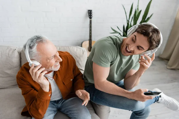High angle view of excited father and son looking at each other while listening music in wireless headphones in living room — Stock Photo