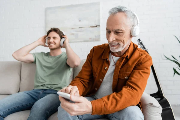 Mature and happy man in wireless headphones using smartphone near son listening music on blurred background — Stock Photo