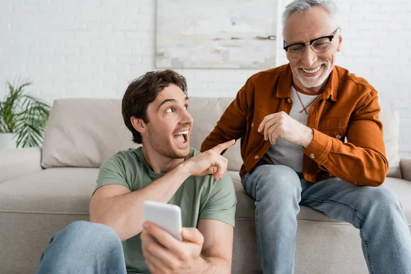 Uomo eccitato che punta a papà maturo allegro mentre tiene il telefono cellulare offuscato — Foto stock