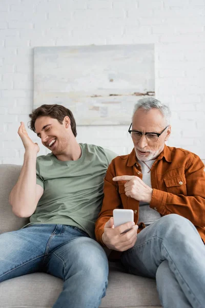 Bärtiger und reifer Mann zeigt bei Videoanruf auf Smartphone mit dem Finger auf lächelnden Sohn — Stockfoto