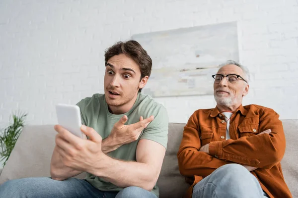 Astonished young man gesturing during video call while sitting near mature father with crossed arms — Stock Photo