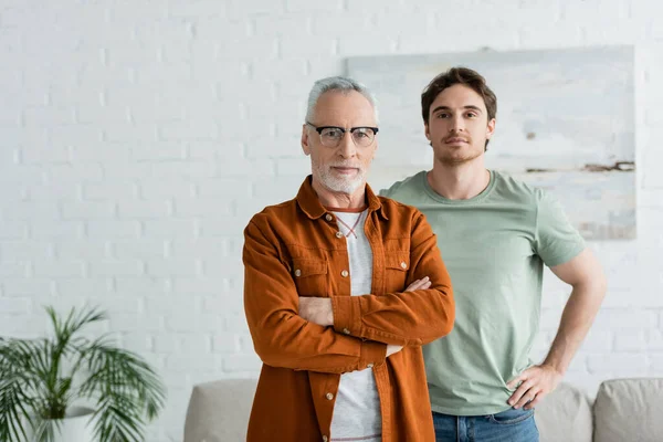 Young man standing with mature father in eyeglasses posing with crossed arms at home — Stock Photo