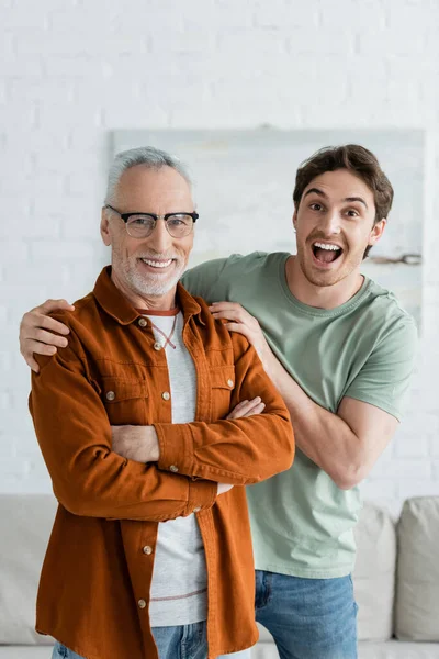 Amazed young man hugging mature father in eyeglasses standing with crossed arms at home — Stock Photo