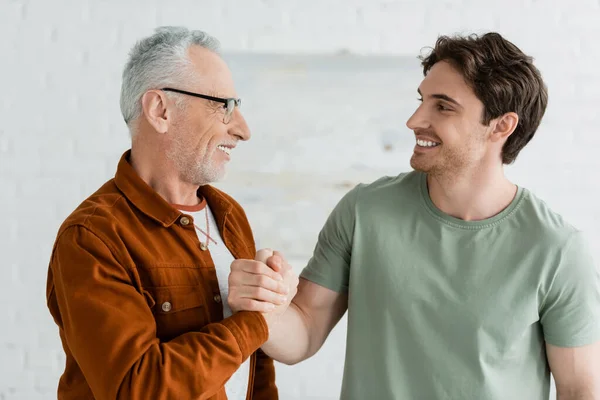 Happy young man shaking hands with cheerful and mature father in eyeglasses at home — Stock Photo