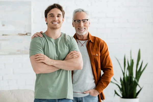 Happy mature father in eyeglasses hugging cheerful young son posing with crossed arms at home — Stock Photo