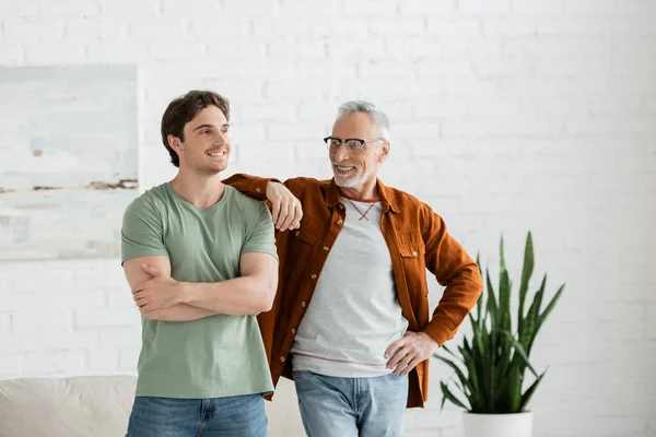 Happy mature father in eyeglasses leaning on shoulder of cheerful son posing with crossed arms at home — Stock Photo