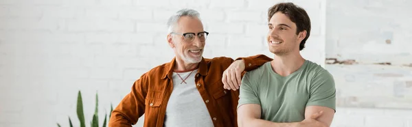 Happy mature father in eyeglasses leaning on shoulder of cheerful young son posing with crossed arms at home, banner — Stock Photo