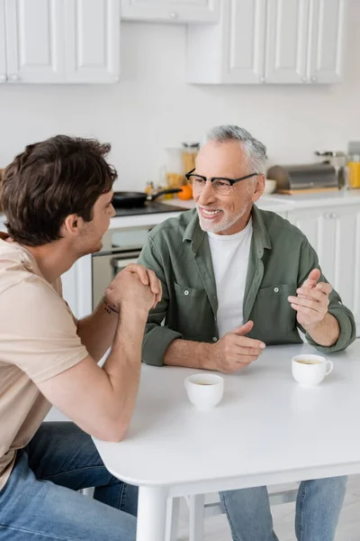 Cheerful and mature father in eyeglasses gesturing while talking with son near cups of coffee — Stock Photo