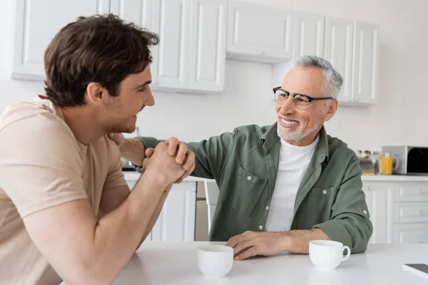 Cheerful and mature father in eyeglasses hugging shoulder of happy son during breakfast — Stock Photo