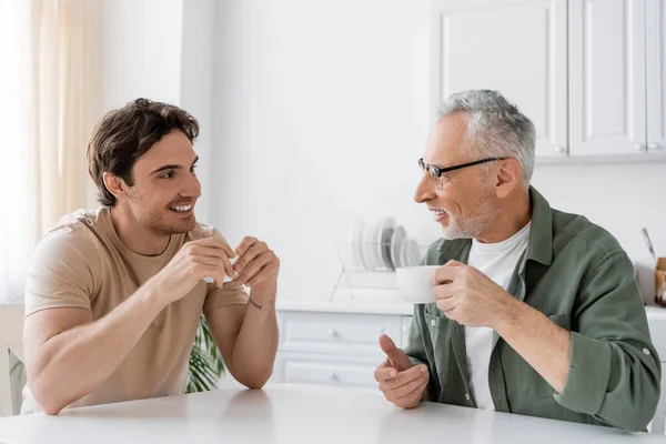 Happy young man drinking morning coffee with smiling and mature father at home — Stock Photo