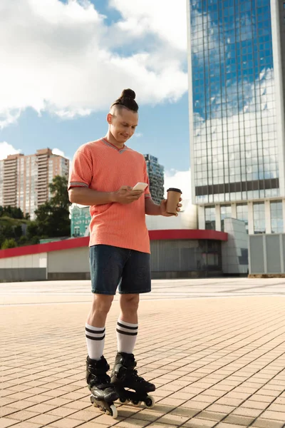 Patinador sonriente usando smartphone y sosteniendo café para salir a la calle urbana - foto de stock