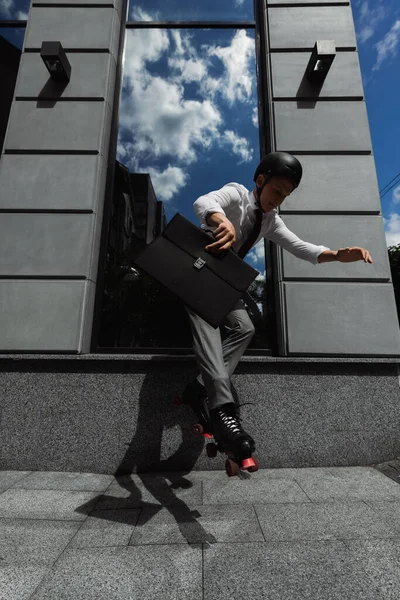 Молодой менеджер в роликовых коньках и формальной одежде держит портфель и прыгает рядом со зданием на открытом воздухе — стоковое фото