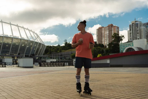Vue latérale du jeune homme en patins à roulettes tenant smartphone et tasse en papier dans la rue urbaine — Photo de stock