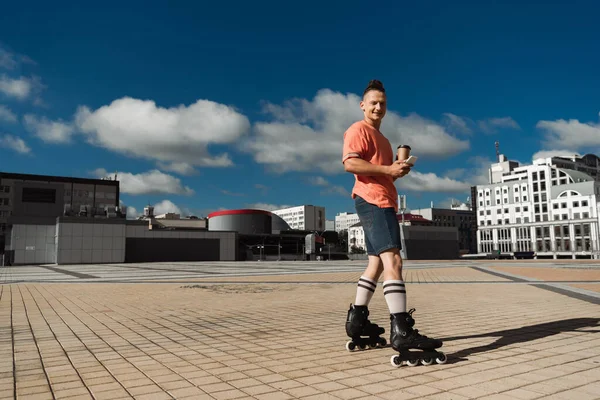 Позитивный роликовый конькобежчик держит сотовый телефон и кофе, чтобы пойти, глядя на камеру на городской улице — стоковое фото