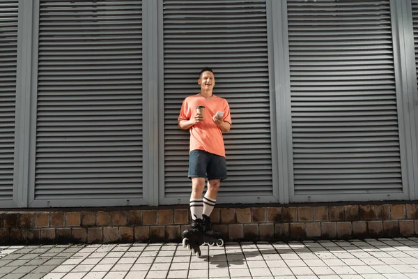 Улыбающийся мужчина в роликовых коньках держит мобильный телефон и кофе, чтобы подойти к зданию на улице в дневное время — стоковое фото