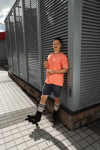 Homme positif en patins à roulettes riant tout en tenant smartphone et café dans la rue urbaine — Photo de stock